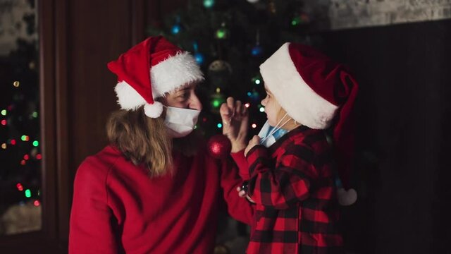 coronavirus, covid-19 in the new year. little daughter with her mother in a bad Santa hat and medical mask under the Christmas tree, remove the masks