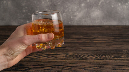 Hand holding glass of whiskey with ice cubes on rustic wooden table with copy-space