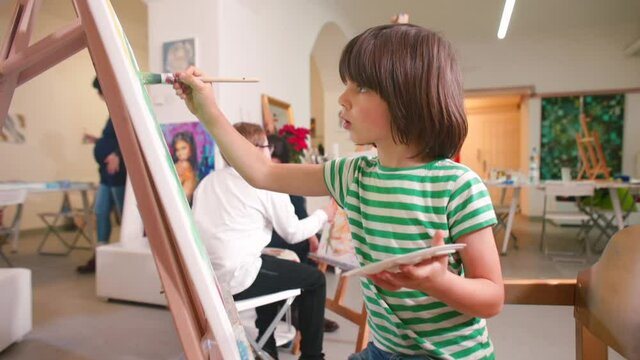The creative atmosphere of an art studio. A cute long-haired boy of elementary school age paints a picture with a brush. Talent identification and development in children.