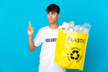 holding a bag full of plastic bottles to recycle over blue background with fingers crossing and...