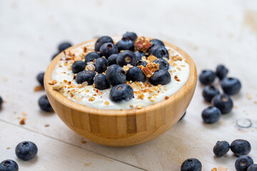 Close up of a wooden bowl filled with white yoghurt and blueberries on white table. Detailed view of healthy nutritious breakfast with fruit and cereals.