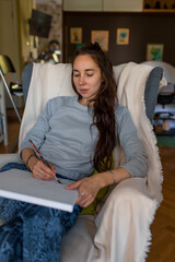 A woman at home in an armchair, paints in her hand a picture of a white frame. Painting background on the wall, long hair. Home studio creativity and art concept.