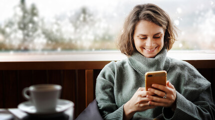 Young woman in a green sweater using a mobile phone by the window in her country house with a view...