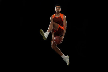 Flying. Young purposeful african-amrican basketball player training, practicing in action, motion isolated on black background. Concept of sport, movement, energy and dynamic, healthy lifestyle.