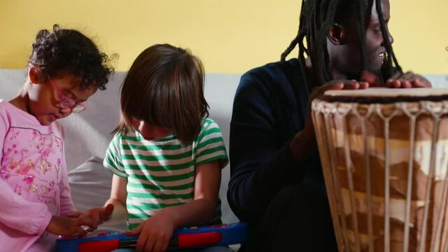 Shows the technique of striking jambe with his hands. Transfer of knowledge to the generation. Training of technique and musical ear. Black father teaches children to play son's drum. 