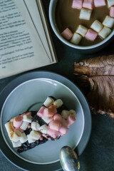 Hot chocolate or cocoa with marshmallows, rocky road brownies on a plate and a book on top of stone grey background