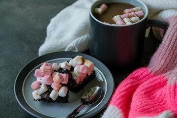Hot chocolate or cocoa with marshmallows and rocky road brownies on a plate over cozy background with colorful warm sweater