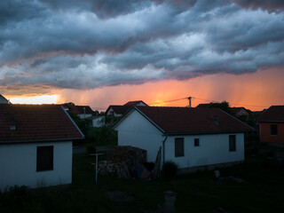 Stormy severe weather with dark clouds, cloudburst or shower and sunshine behind rain above settlement during summer evening.