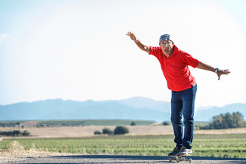Uomo settantenne con polo rossa, va nel suo skateboard stando in equilibrio in una strada di...