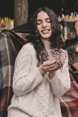 A woman of model appearance with long dark curly hair in a white sweater sits on a bench and holds a New Year's garland in her hands