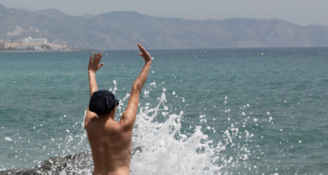Mujer feliz de vacaciones en la playa sola, feliz jugando con olas que le salpican al chocar en las rocas con un pa&ntilde;uelo en la cabeza, vistas de atr&aacute;s