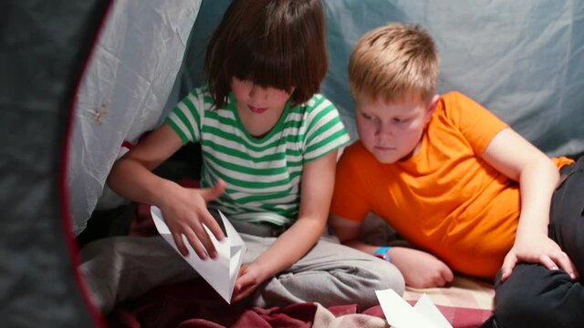 Two children in a tent are making origami paper. Fold the paper into a shape. Useful self-isolation entertainment.