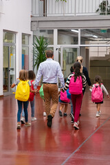 Schoolkids with bright backpacks walking through school hallway, holding hands of teachers. Back view, full length. Education or back to school concept