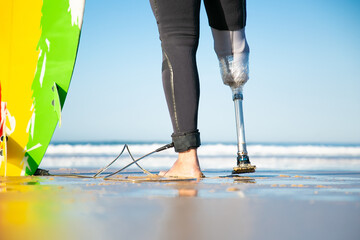 Closeup view of artificial leg of disabled surfer standing near surfboard. Feet of male amputee on sea coast. Ocean waves on background. Physical disability, lifestyle and extreme sport concept