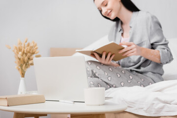 laptop, cup and book on coffee table near cheerful woman with vitiligo on blurred background