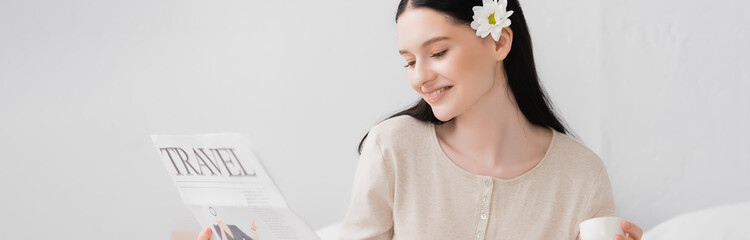 Fototapeta premium cheerful woman with flower in hair holding cup and reading travel newspaper, banner