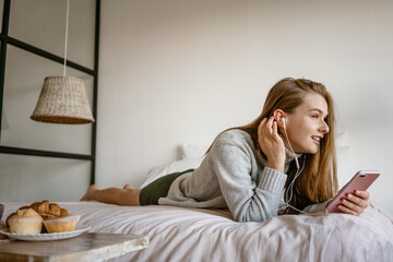 Beautiful smiling girl using cellphone and earphones while lying on bed