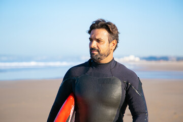 Portrait of handsome surfer standing on beach with surfboard and looking away. Pensive Caucasian middle-aged man holding surfboard and thinking about something. Lifestyle and summer activity concept