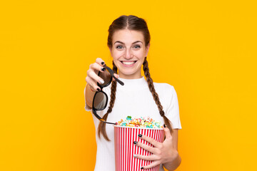 Young woman over isolated yellow background with 3d glasses and holding a big bucket of popcorns