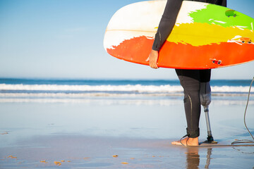 Back view of cropped male surfer standing with surfboard on sea beach. Unrecognizable amputee with...