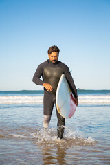Pensive handicapped surfer walking in sea water with board. Bearded middle-aged amputee with artificial leg carrying surfboard and looking down. Physical disability and extreme sport concept