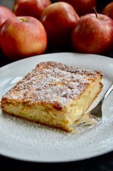 One piece of a traditional german apple cake on a white plate with fork, powdered sugar on top, blurred apples in the background
