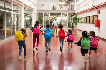 Group of schoolkids wearing bright backpacks, running to favorite teacher through school hallway....