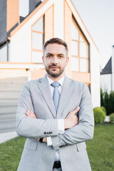 Front view of confident broker with crossed arms looking at camera with blurred house on background