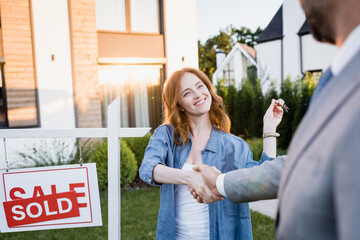 Happy redhead woman with keys shaking hands with broker near sign with sold lettering on blurred...