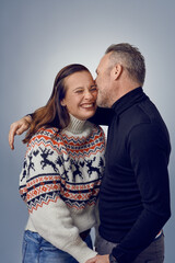 Adult man kissing his happy laughing wife. Loving couple in warm sweaters posing for family portrait. Winter holidays photoshoot concept in studio