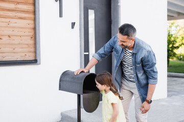 Smiling father standing near daughter looking in empty mailbox near house