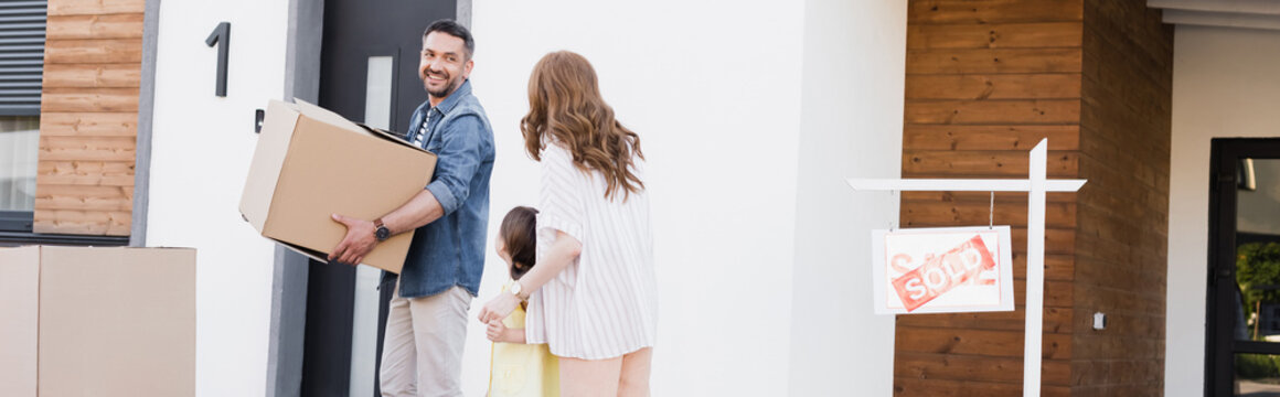 Smiling Husband With Carton Box Looking At Wife Holding Hands With Daughter Near House And Sign With Sold Lettering, Banner