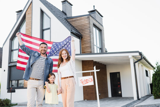 Cheerful couple with daughter holding american flag while looking at camera near house and sign with sold lettering