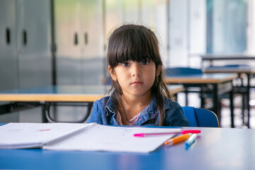 Serious Latin girl sitting at school desk and looking at camera. Front view. Education or back to school concept