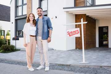 Full length of happy couple looking at camera and hugging while standing near sign with sold lettering and house