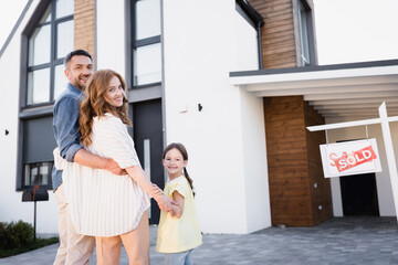 Happy family with daughter hugging and looking at camera near sign with sold lettering and house - obrazy, fototapety, plakaty