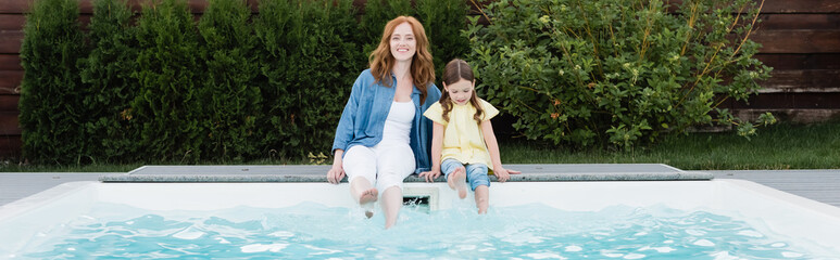 Fototapeta na wymiar Cheerful redhead mother looking at camera while sitting with daughter near swimming pool on backyard, banner