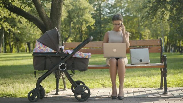 Wide shot of concentrated young businesswoman sitting in summer park with baby stroller, laptop, and talking on the phone. Portrait of confident busy Caucasian woman working and taking care of infant.