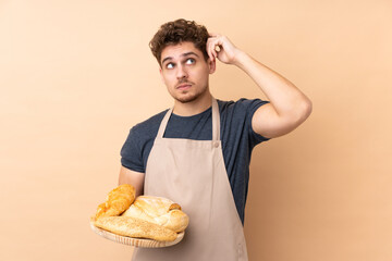 Male baker holding a table with several breads isolated on beige background having doubts and with confuse face expression