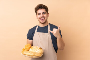 Male baker holding a table with several breads isolated on beige background making phone gesture