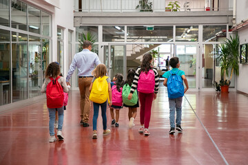 Schoolkids with colorful backpacks and male teacher walking through school hallway. Back view, full...