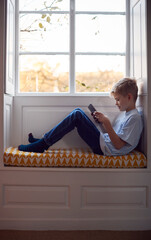 Young Boy Sitting On Window Seat At Home Playing On Digital Tablet