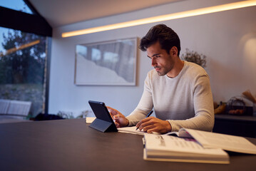 Evening Shot Of Man In Kitchen Working Or Studying From Home Using Digital Tablet