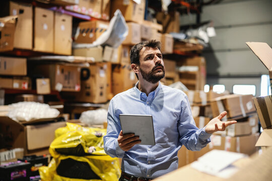 Confused chief standing in storage full of boxes ready for shipment and holding tablet. He is checking on goods. Export firm.