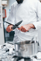 Chef in white uniform standing at kitchen. Holding knives in hands