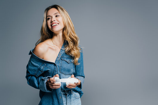 happy sensual woman in denim shirt, with naker shoulder, holding wireless headphones isolated on grey