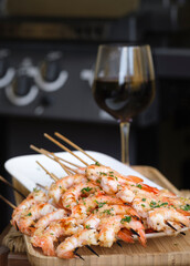 Grilled king prawns on skewers and glass of red wine. Background is a gas grill in bokeh