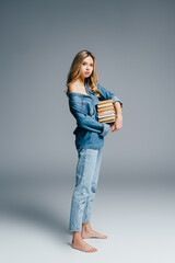 sexy barefoot woman in denim clothes, with naked shoulder, holding stack of books on grey