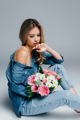 seductive young woman, wearing denim clothes, sitting with naked shoulder and holding flowers on grey