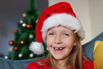 Cute blonde caucasian girl 7 years old sitting on couch near Christmas tree and writing letter to Santa Claus. Bedroom decorated with garlands. Xmas celebrate, happy new year 2021. Time to make wishes
